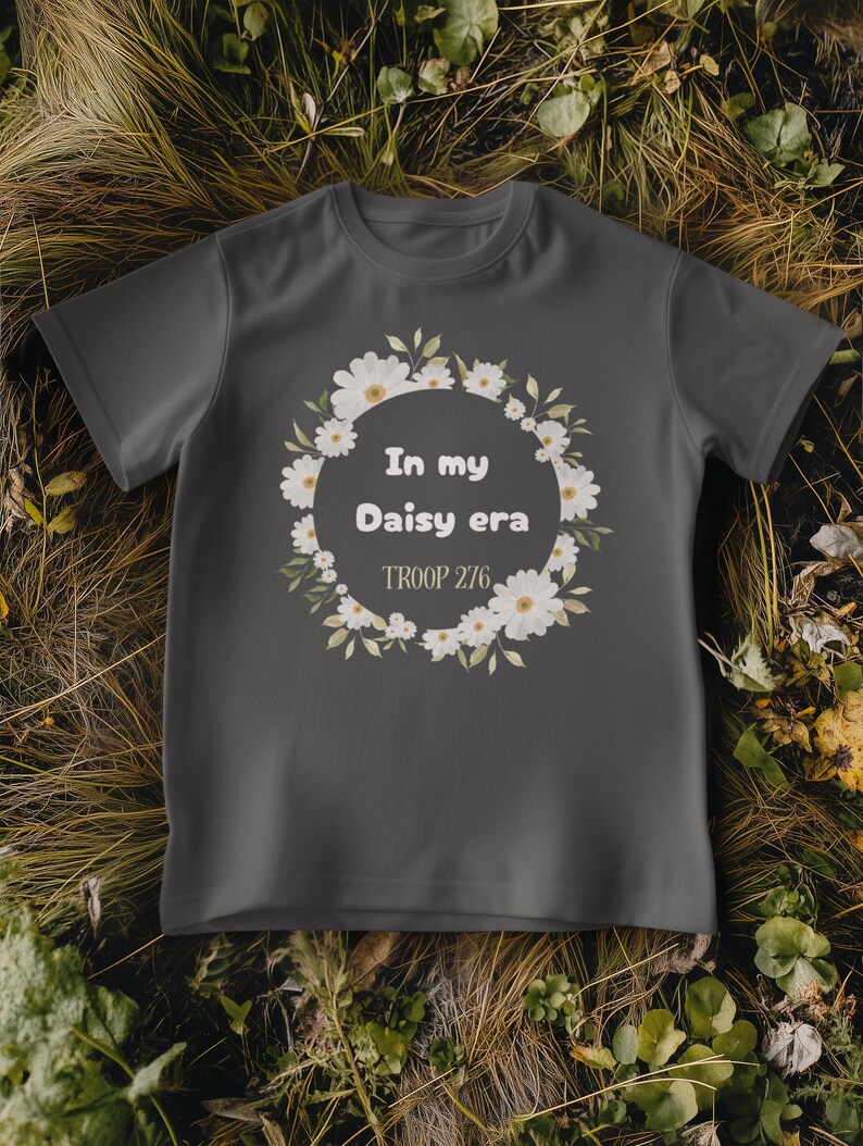 Op de afbeelding: Een donkergrijs t-shirt met een bloemenkransontwerp van witte madeliefjes en groene bladeren. De tekst "In my Daisy era" en "TROOP 276" staan in de krans. Het shirt wordt gepresenteerd op droog gras en bladeren.