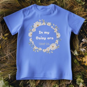 Op de afbeelding: Een lichtblauw T-shirt met de tekst "In my Daisy era" in een krans van witte madeliefjes en groene bladeren. De tekst "TROOP 276" staat eronder. Het T-shirt wordt getoond op een bed van droog gras en bladeren.