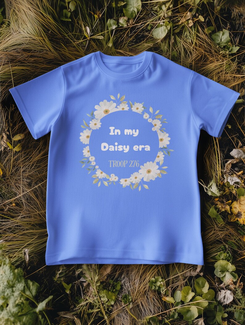 Op de afbeelding: Een lichtblauw T-shirt met de tekst "In my Daisy era" in een krans van witte madeliefjes en groene bladeren. De tekst "TROOP 276" staat eronder. Het T-shirt wordt getoond op een bed van droog gras en bladeren.