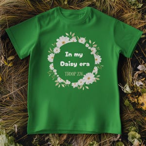 Op de afbeelding: Een groen t-shirt met de tekst "In my Daisy era" in een bloemenkrans. De tekst "TROOP 276" staat eronder. Het t-shirt wordt gepresenteerd op een bed van droog gras en bladeren.