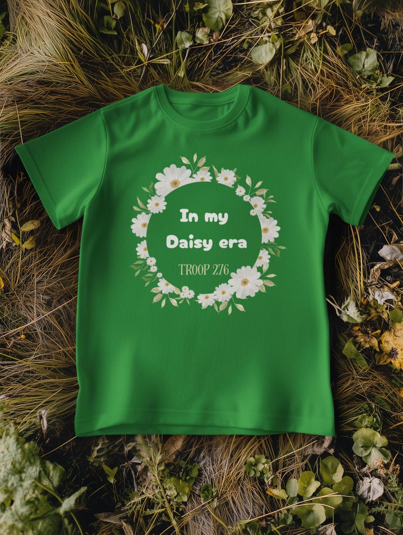 Op de afbeelding: Een groen t-shirt met de tekst "In my Daisy era" in een bloemenkrans. De tekst "TROOP 276" staat eronder. Het t-shirt wordt gepresenteerd op een bed van droog gras en bladeren.