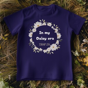 Op de afbeelding: Een marineblauw t-shirt met de tekst "In my Daisy era" in een krans van witte madeliefjes. De tekst "TROOP 276" staat eronder. Het t-shirt wordt getoond op een bed van droog gras en bladeren.