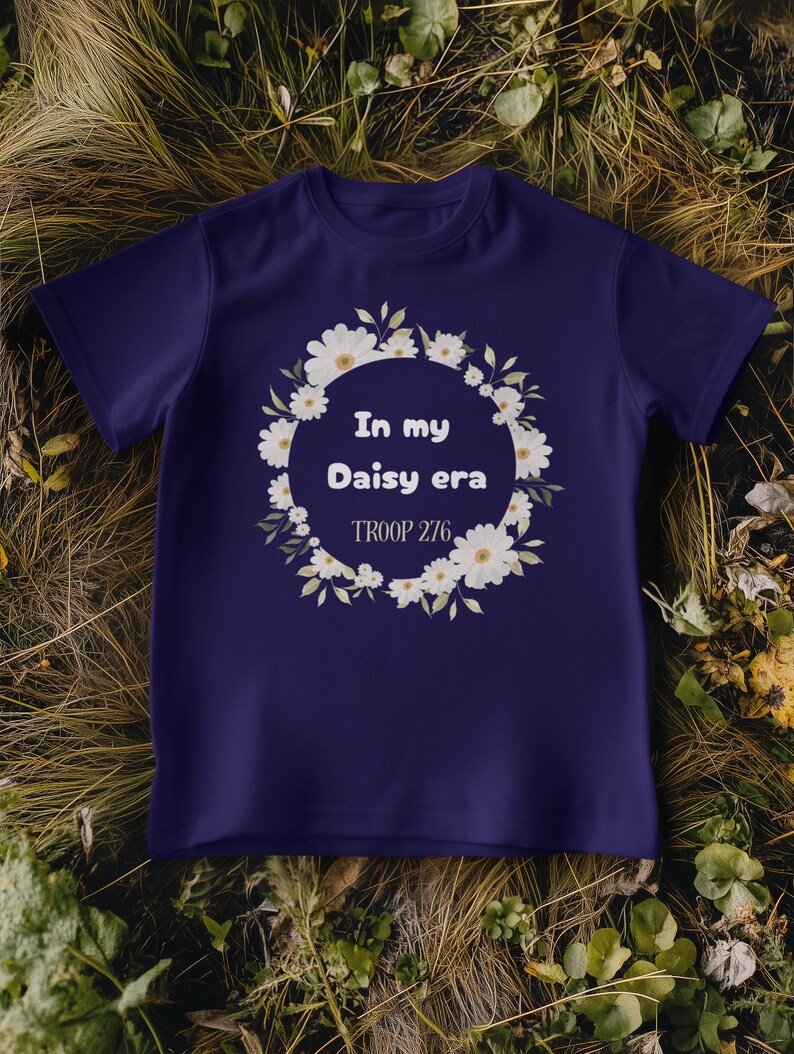 Op de afbeelding: Een marineblauw t-shirt met de tekst "In my Daisy era" in een krans van witte madeliefjes. De tekst "TROOP 276" staat eronder. Het t-shirt wordt getoond op een bed van droog gras en bladeren.