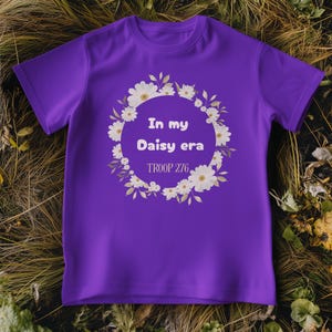 Op de afbeelding: Een paars T-shirt met de tekst "In my Daisy era" in een bloemenkrans. De tekst "TROOP 276" staat eronder. Het shirt is gemaakt van zacht materiaal en heeft korte mouwen.