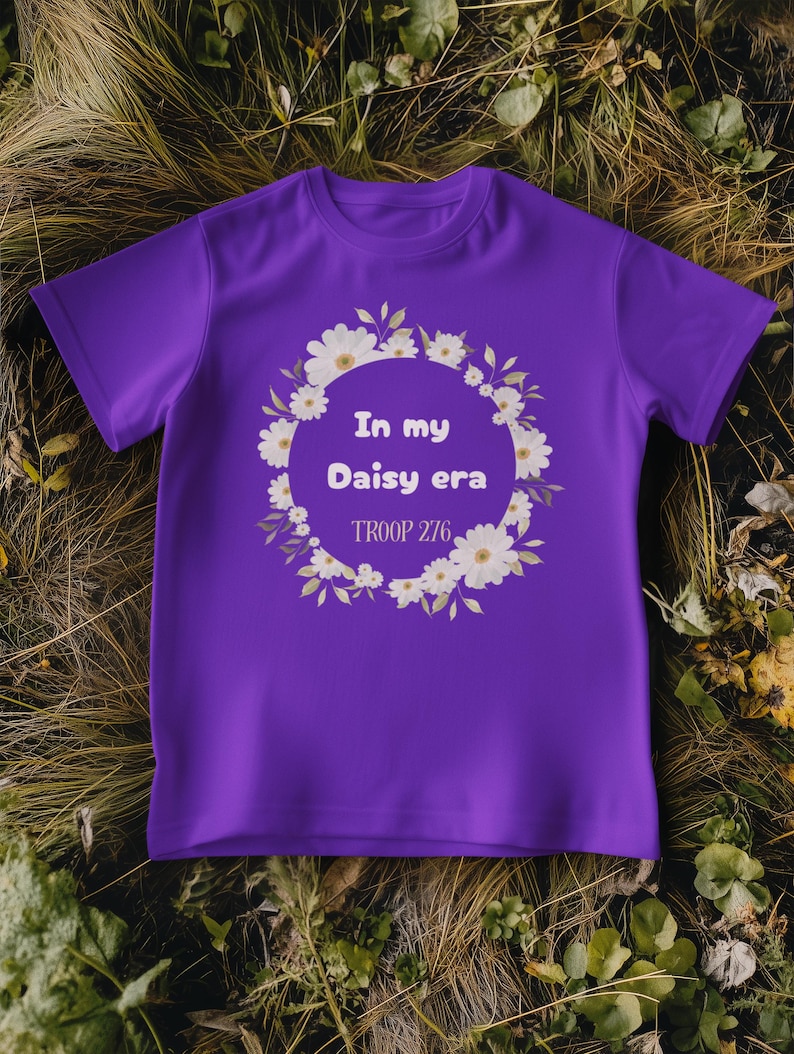 Op de afbeelding: Een paars T-shirt met de tekst "In my Daisy era" in een bloemenkrans. De tekst "TROOP 276" staat eronder. Het shirt is gemaakt van zacht materiaal en heeft korte mouwen.