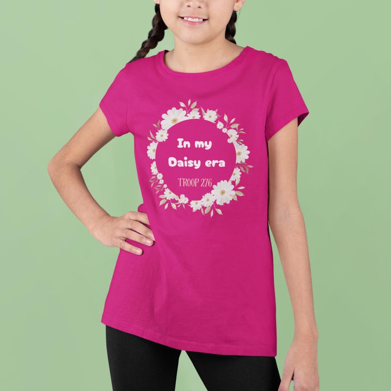 Op de afbeelding: Een magenta T-shirt met de tekst "In my Daisy era" in een bloemenkrans, en "TROOP 276" eronder. Het shirt heeft een effen magenta kleur.