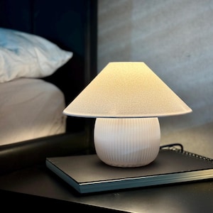 Könnte beinhalten: Eine kleine, weiße Nachttischlampe mit konischem Schirm und geripptem Sockel. Die Lampe steht auf einer schwarzen Oberfläche, neben einem Notizbuch und einem Glas. Der Lampenschirm ist cremefarben, der Sockel gerillt.