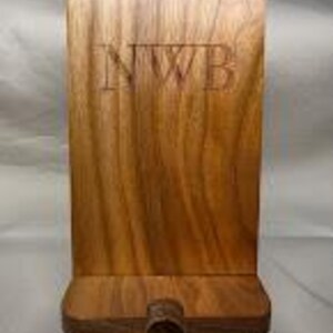 May include: A wooden phone stand with a rectangular back and a small base. The stand is made of a light brown wood and has the letters "NWB" carved into the front. The stand has a slot at the bottom to hold a phone.