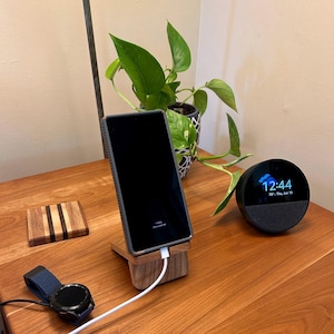 May include: A wooden phone stand with a charging phone, a smart watch, a coaster, and a smart speaker on a wooden table. A green plant in a decorative pot is in the background. The phone is charging with a white cable.