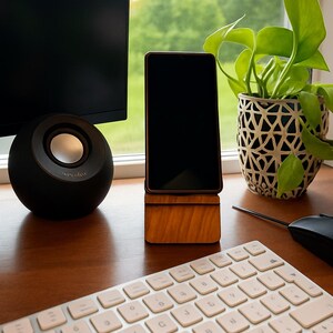 May include: A wooden phone stand with a phone in it, a black speaker, a white keyboard, and a green plant in a decorative pot sit on a wooden desk. A computer monitor is visible in the background.