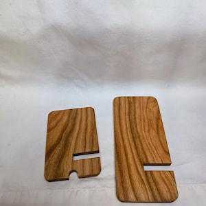 May include: Two wooden phone stands with a natural wood grain finish. Each stand has a rectangular shape with a slot for holding a phone. One stand has a rounded bottom edge. The stands are displayed on a white background.