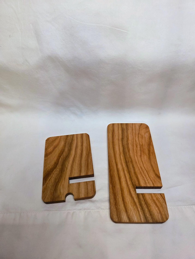 May include: Two wooden phone stands with a natural wood grain finish. Each stand has a rectangular shape with a slot for holding a phone. One stand has a rounded bottom edge. The stands are displayed on a white background.