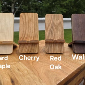 May include: Four wooden phone stands in different wood types: Hard Maple, Cherry, Red Oak, and Walnut. Each stand has a rectangular shape with a slot to hold a phone. The wood grains and colors vary, with text labels below each stand.