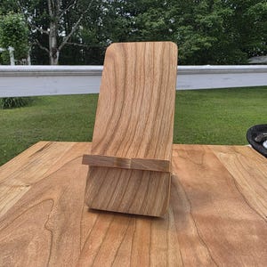 May include: A wooden phone stand with a natural wood grain finish. The stand has a rectangular base and a back support with a small ledge to hold a phone or tablet. The stand is made of light brown wood and sits on a wooden surface.