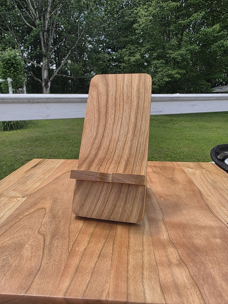 May include: A wooden phone stand with a natural wood grain finish. The stand has a rectangular base and a back support with a small ledge to hold a phone or tablet. The stand is made of light brown wood and sits on a wooden surface.