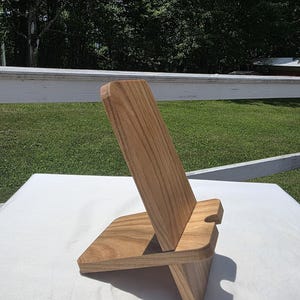 May include: A wooden phone stand with a natural wood grain finish. The stand has a rectangular base and a slanted back support with a slot for holding a phone. The design is simple and modern.
