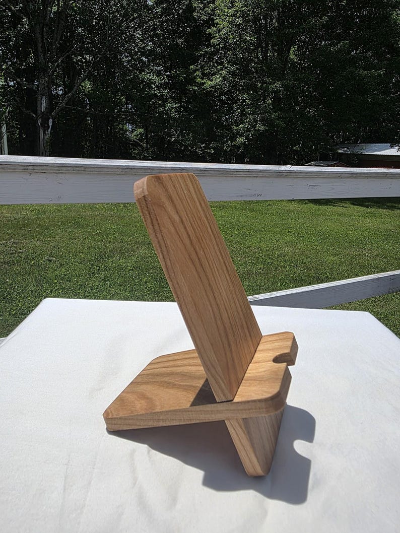 May include: A wooden phone stand with a natural wood grain finish. The stand has a rectangular base and a slanted back support with a slot for holding a phone. The design is simple and modern.