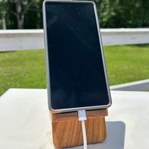 May include: A smartphone with a black screen is propped up on a wooden phone stand. The stand is made of light brown wood and has a slot to hold the phone. A white charging cable is plugged into the phone.
