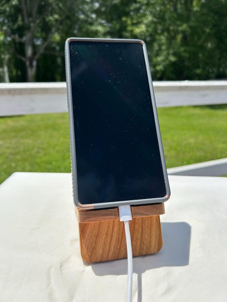 May include: A smartphone with a black screen is propped up on a wooden phone stand. The stand is made of light brown wood and has a slot to hold the phone. A white charging cable is plugged into the phone.