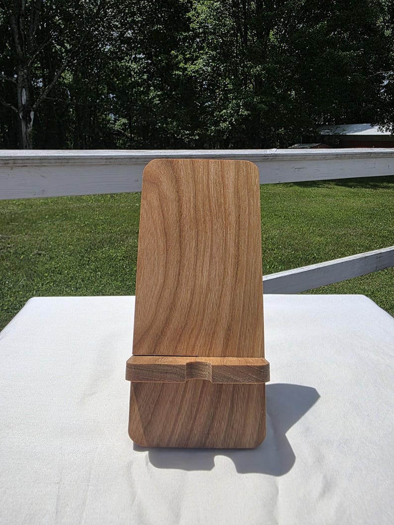 May include: A wooden phone stand with a natural wood grain finish. The stand has a rectangular base and a tall, slightly curved back. A small ledge is carved into the base to hold a phone. The stand is made of light brown wood.