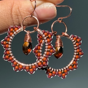 May include: Handmade copper earrings with a floral design. The earrings feature a circular frame adorned with orange and burgundy beads, and a dangling faceted amber-colored crystal. The earrings are suspended from copper ear wires.