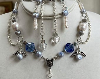May include: A delicate necklace and earring set featuring silver-toned chains, angel wing charms, and a mix of blue, gray, and white pearls. The necklace includes blue square beads and a heart-shaped pendant. The earrings have dangling pearl and crystal accents.