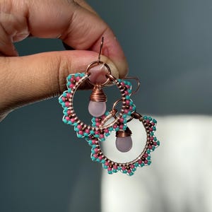 May include: A pair of handmade hoop earrings. The earrings feature a copper-colored metal frame, adorned with turquoise and pink beads. Each earring has a teardrop-shaped, light pink gemstone pendant and copper wire detailing.