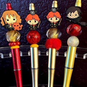 May include: Four decorative pens featuring cartoon character toppers. The pens have red, gold, and yellow barrels. The toppers include characters with red hair, glasses, and a cat. The pens are displayed on a clear stand.