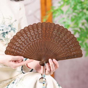 May include: A brown wooden hand fan with intricate carved designs and a decorative tassel. The fan is held open, showcasing its detailed craftsmanship. The fan's design features a repeating pattern of swirls and lines.