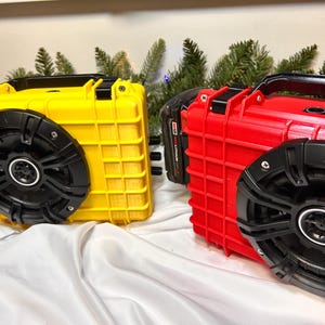 May include: Two portable speakers, one yellow and one red, with black speaker grilles and handles. The speakers have a rugged, waterproof design. The image shows the speakers on a white surface with evergreen branches in the background.