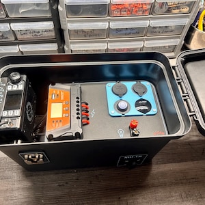 May include: Black portable power box with a radio, orange control panel, and blue panel with outlets. The box is open, revealing the internal components. The box has a latch and the text "SOLAR" on the front.
