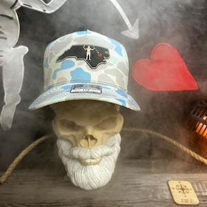 May include: A camouflage-patterned baseball cap with a North Carolina outline patch featuring a pirate design. The hat is displayed on a skull with a white beard. A red heart and a compass are also visible.