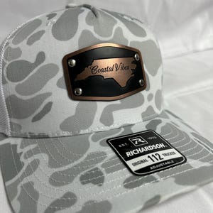 May include: A gray and white camouflage trucker hat with a white mesh back. The hat features a brown leather patch with the words "Coastal Vibes" and a North Carolina outline. A black and white tag reads "Richardson 112 Trucker".