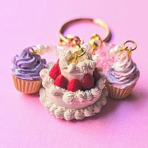 Puede incluir: Un llavero dorado con dijes de pastel y cupcake en miniatura. Los dijes incluyen un pastel de dos niveles con bayas rojas y dos cupcakes con glaseado morado y plateado. El fondo es de color rosa liso.