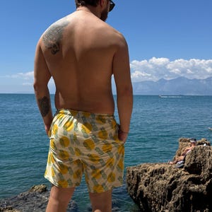May include: Yellow and white pineapple print swim trunks. The shorts are knee-length and worn by a person standing on a rocky shore, with the ocean and mountains in the background. The person is wearing white sneakers.
