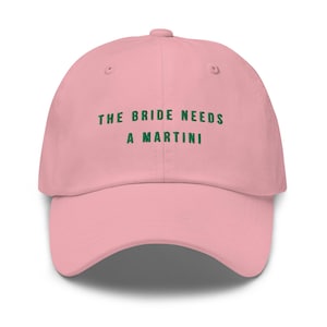 May include: A light pink baseball cap with the text "THE BRIDE NEEDS A MARTINI" in green lettering. The cap has a curved brim and a fabric strap closure on the back. A fun accessory for a bachelorette party or bridal event.