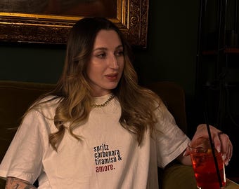 Könnte beinhalten: Ein weißes T-Shirt mit den Worten "spritz, carbonara, tiramisu & amore" in rotem Text. Die Person hält ein Glas mit roter Flüssigkeit mit einem Strohhalm. Die Person sitzt auf einem grünen Samtsofa.