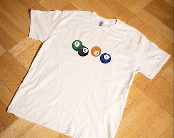 May include: A cream-coloured t-shirt with the word "POOL" spelled out using billiard balls. The balls are green, black, orange, and blue. The shirt has the text "LOOM FOLK APPAREL CO." in the upper right corner.