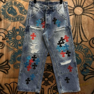 Könnte beinhalten: Hellblaue Jeans mit Distressed-Details und gestickten Kreuzmustern in Rot, Schwarz und Blau. Die Jeans hat einen Knopfverschluss und mehrere Taschen. Der Stil ist lässig und edgy.