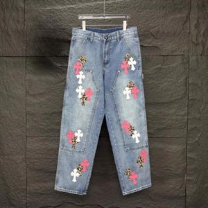 May include: Light blue denim jeans with multiple cross and leopard print patches. The crosses are white and pink. The jeans are hanging on a silver hanger.