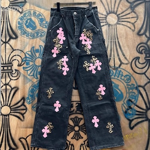 May include: Black denim trousers with pink cross and leopard print patches. The trousers have a classic five-pocket design and a button and zip closure. The trousers are hanging on a black hanger.