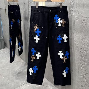 May include: Black denim jeans featuring multiple cross patches in white, blue, and leopard print. The jeans are displayed on a hanger, highlighting the unique design. A second pair of jeans is visible in the background.