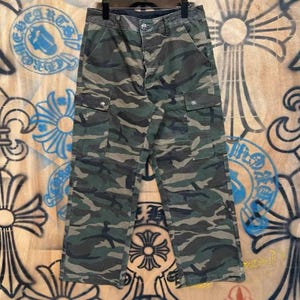 May include: Camouflage cargo pants in shades of green, brown, and black. The pants feature multiple pockets and a button closure. The fabric has a worn appearance. The pants are hanging on a black hanger.