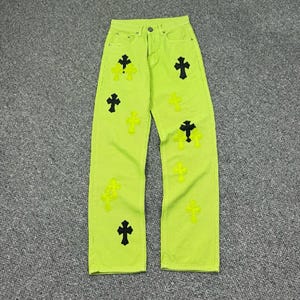 May include: Vibrant neon green denim jeans adorned with black and neon yellow cross appliqués. These jeans feature a classic five-pocket design and a straight leg cut. The crosses are of different sizes and are spread across the front of the jeans.