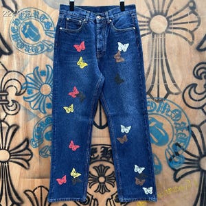May include: Dark blue denim jeans decorated with colourful butterfly patches. The jeans have a classic five-pocket design and a button-fly closure. The butterflies are in shades of red, yellow, white, brown, and black.