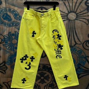 May include: Vibrant yellow denim jeans adorned with black and yellow cross and text details. The word "Chrome" is written in a stylized font on the right leg. The left leg features the number "3" and several cross designs.