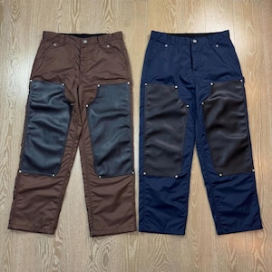 Puede incluir: Dos pantalones de trabajo. Uno es marrón, el otro azul marino. Ambos tienen parches negros en las rodillas y están abotonados en la cintura. Los pantalones están hechos de un material duradero.