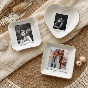 May include: Three white ceramic trinket dishes with gold-toned rims. One heart-shaped dish features a black and white photo and the text I Love You Mom. Two square dishes feature photos and the text I Love You Mom and We love you Mommy.