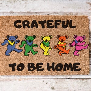 May include: A rectangular doormat made of natural fibers. The mat features the words "GRATEFUL TO BE HOME" in black block letters. Five colorful dancing bear figures are also displayed on the mat, in blue, green, yellow, orange, and pink.