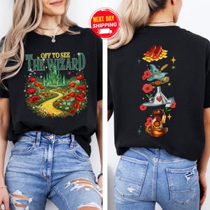 May include: Black t-shirt featuring a colorful graphic from The Wizard of Oz. The front design shows the text "OFF TO SEE THE WIZARD" with a yellow brick road, poppies, and the Emerald City. The back has iconic imagery.